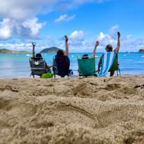 Three friends in beach chairs with arms raised, facing turquoise Caribbean water