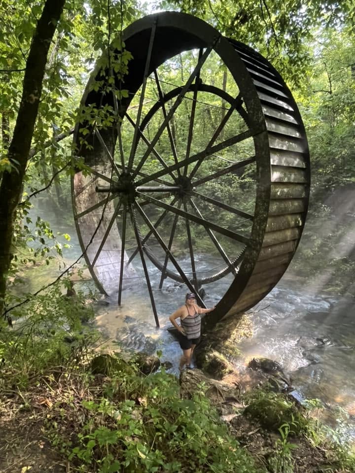A traveler standing beside an enormous waterwheel hidden deep in the forest